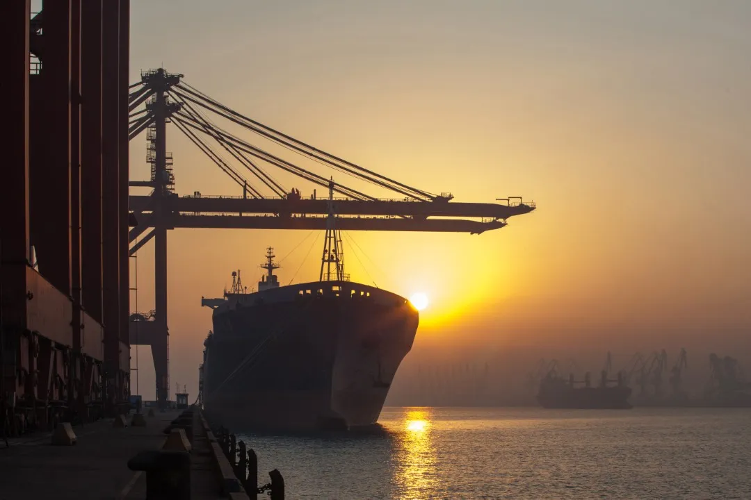 Fotografía de un barco llegando al puerto con el ocaso de fondo y una grúa para descargar el servicio de transporte marítimo